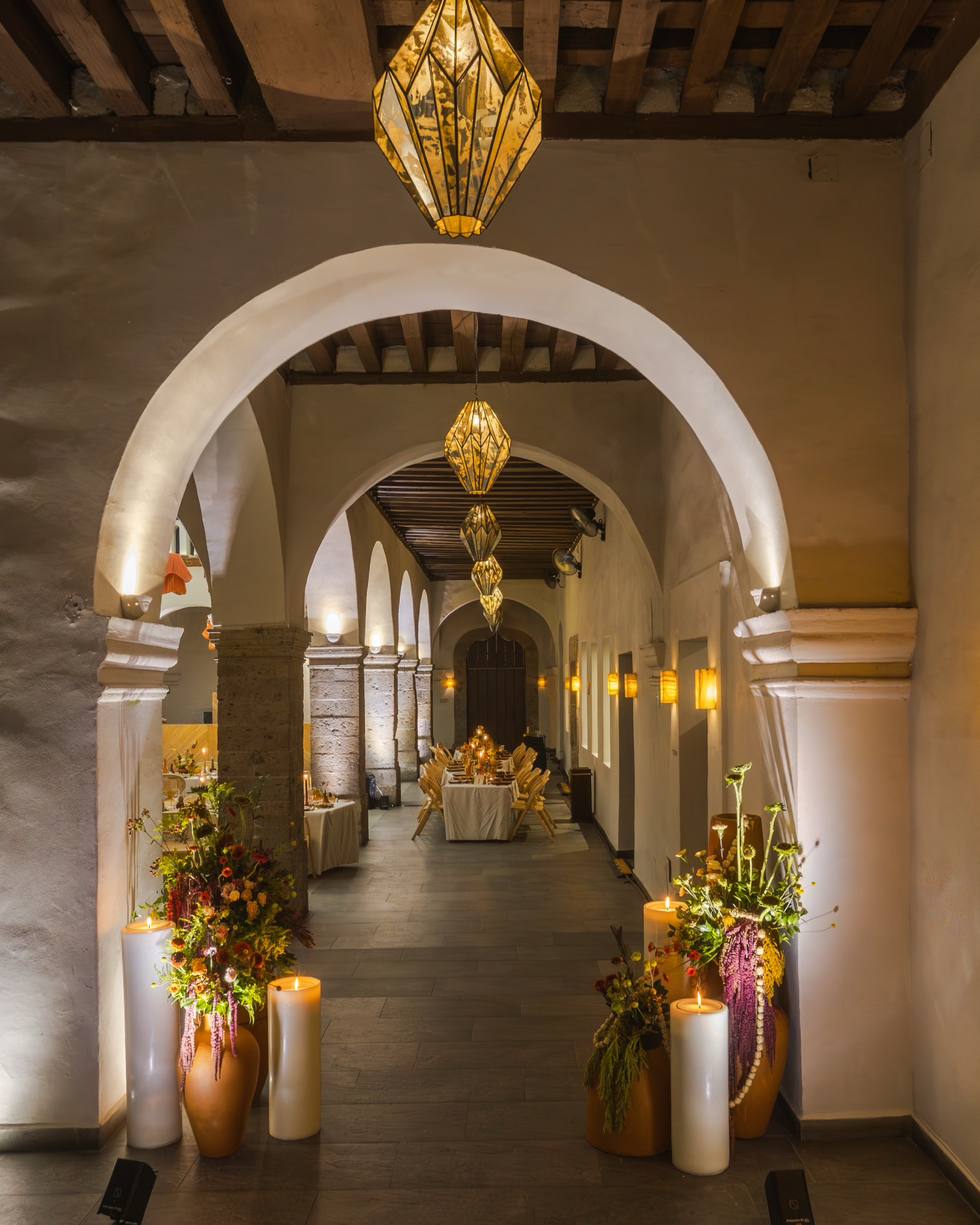 Corredor del Ex Convento visto desde un arco, con florales otoñales, velas y lámparas doradas
